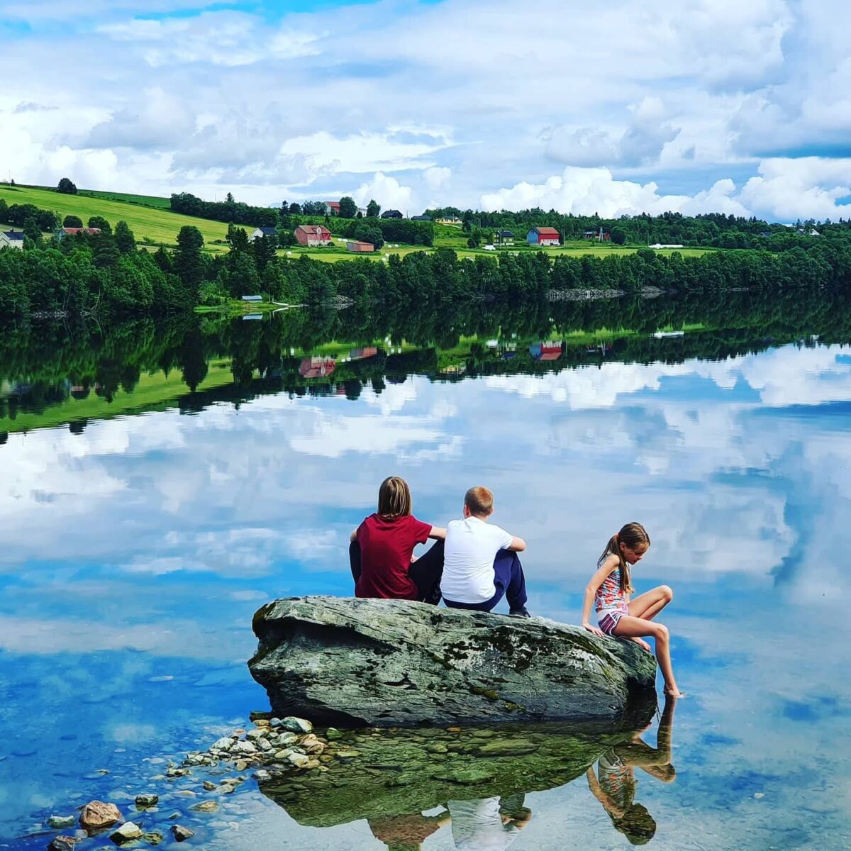 Tre barn sitter på en stor stein ved et vann med gårder i bakgrunnen.