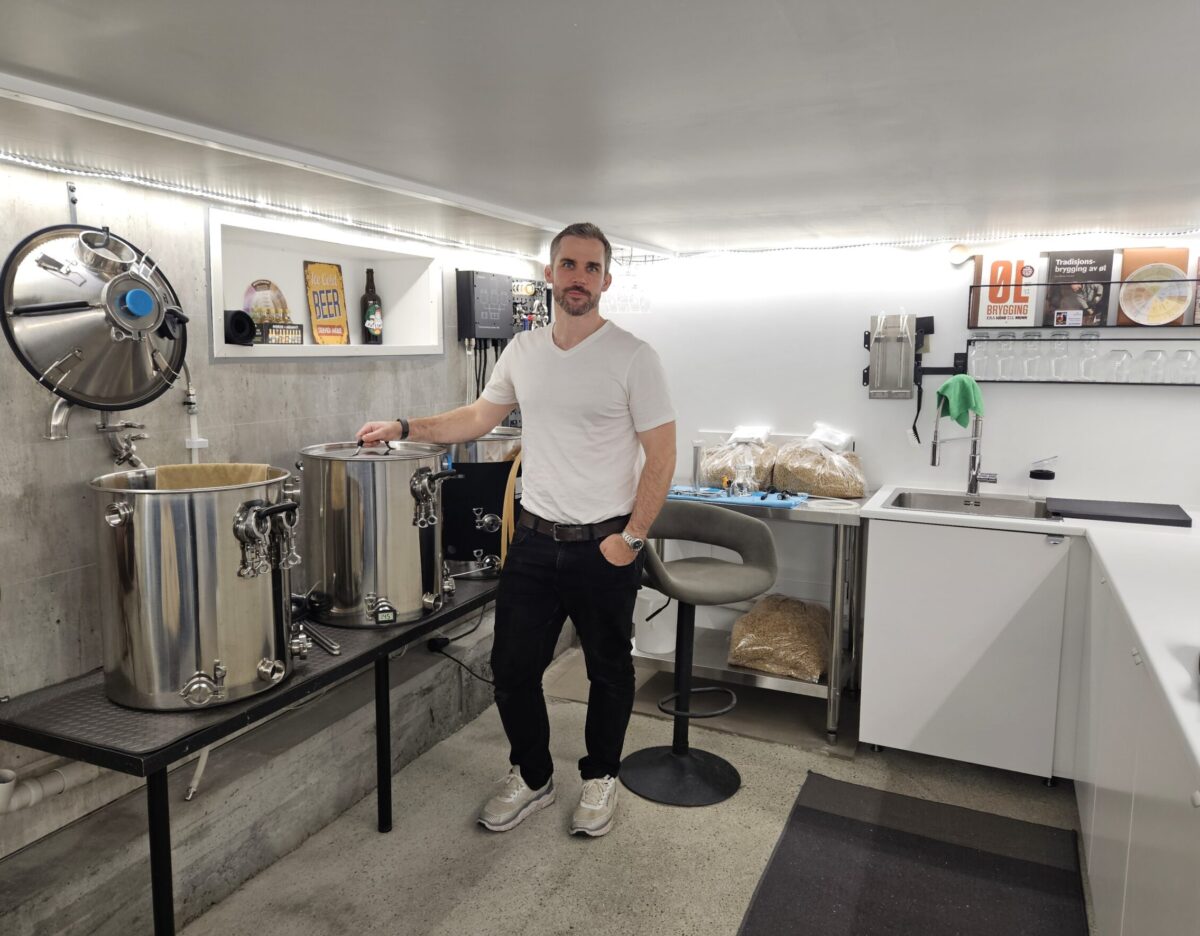 Mann i hvit t-skjorte foran bryggeriutstyr i en kjeller
